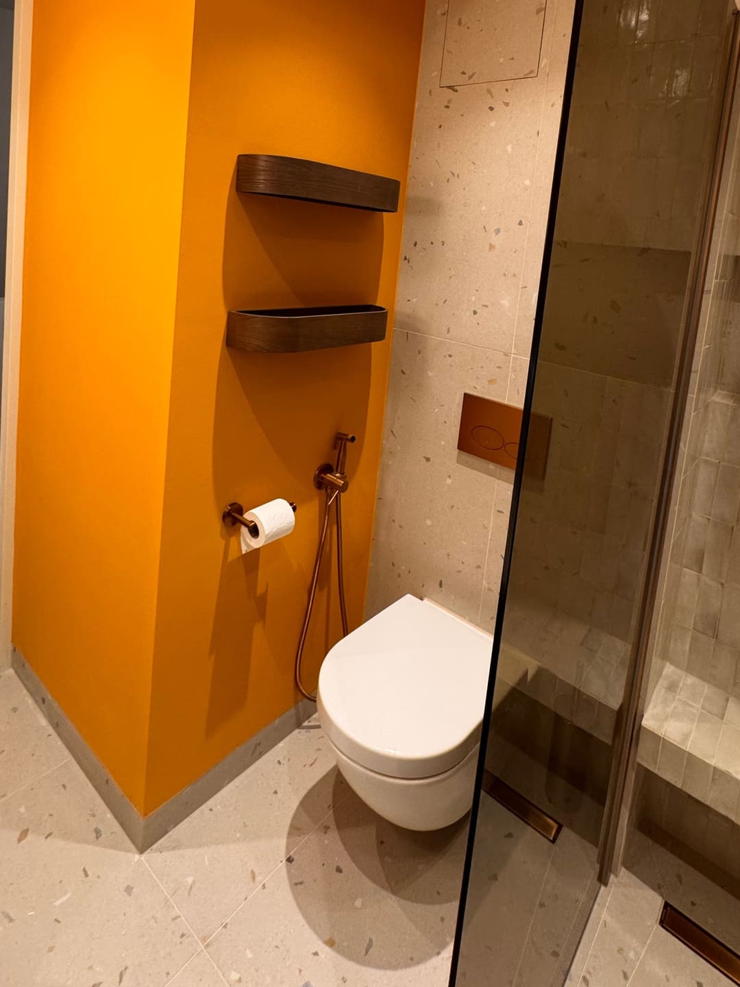 Hangtoilet met koperen bedieningsplaat, terrazzo tegels en een okergele wand met houten planchetten